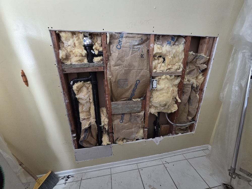 Kitchen wall opened for plumbing repair with exposed studs and insulation during drywall demolition.